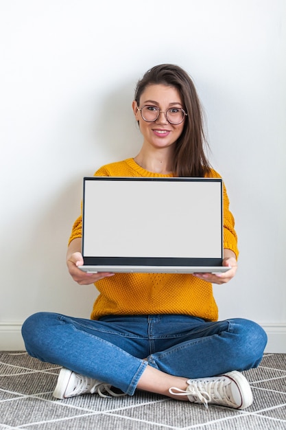 La giovane donna tiene lo schermo in bianco del modello del computer portatile nelle mani, seduto sul tappeto su sfondo bianco