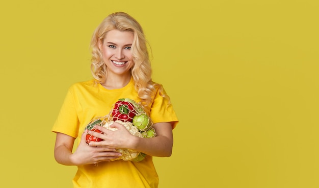 Young woman hold a string bag full of organic vegetables on yellow wall.