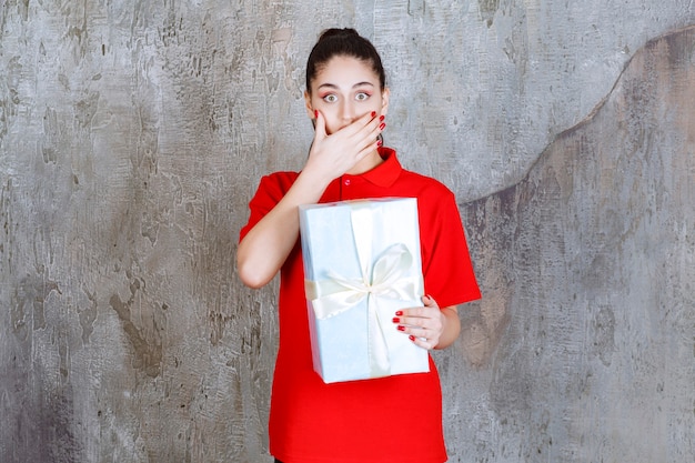 Фото Молодая женщина, держащая синюю подарочную коробку, обернутую белой лентой, выглядит напряженной или напуганной.