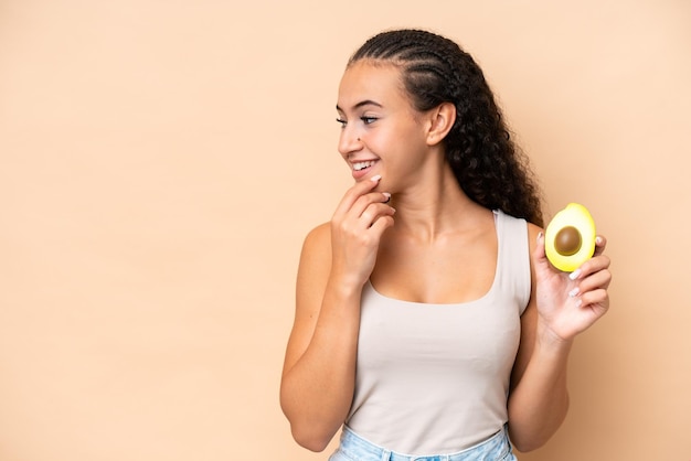 Young woman holding an avocado isolated on beige background thinking an idea and looking side