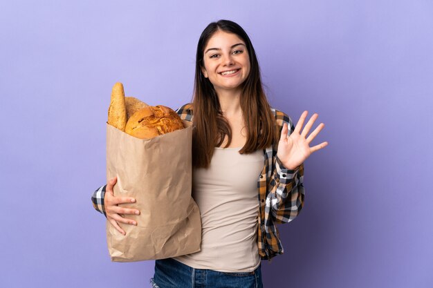 Giovane donna che giudica una borsa piena di pani isolati sul saluto porpora con la mano con l'espressione felice