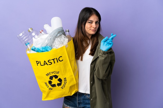 Молодая женщина, держащая сумку, полную пластиковых бутылок для переработки, изолирована на фиолетовом фоне со скрещенными пальцами и желает всего наилучшего