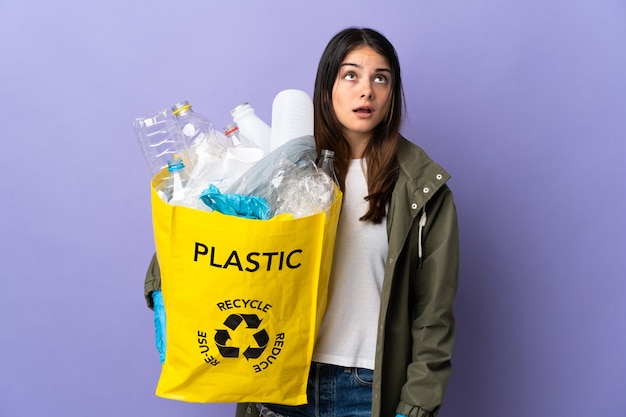 Молодая женщина, держащая сумку, полную пластиковых бутылок для переработки, изолирована на фиолетовой стене, глядя вверх и с удивленным выражением лица