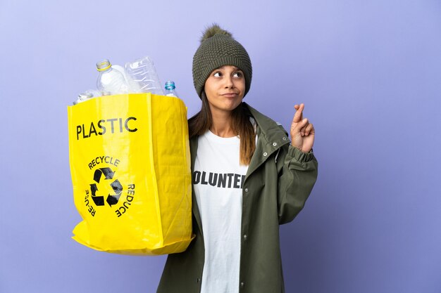 Young woman holding a bag full of plastic with fingers crossing and wishing the best