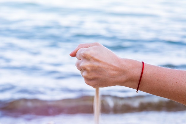 Giovane donna che tiene la sabbia della spiaggia in mano e lascia cadere la sabbia. il vento del mare disperde la sabbia dalla presa della ragazza. sfondo sfocato con onde del mare.