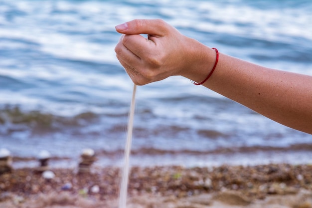 Young woman holding beach sand in the hand and lets sand falling down. Sea wind scatters sand out of girl's grasp. blurred background with sea waves.