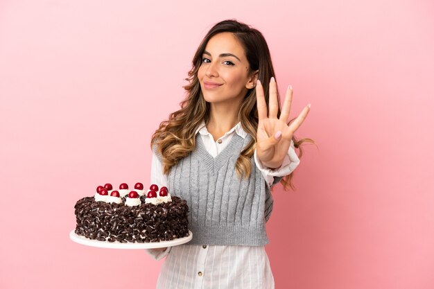 幸せな孤立したピンクの背景の上にバースデーケーキを保持し、指で4を数える若い女性