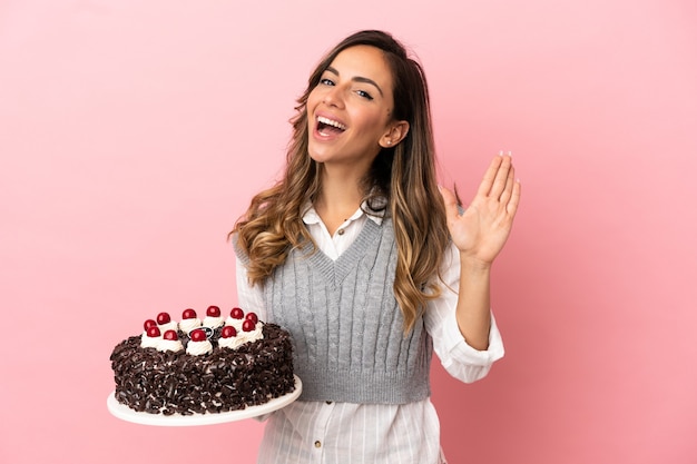 幸せな表情で手で敬礼する孤立したピンクの背景にバースデーケーキを保持している若い女性