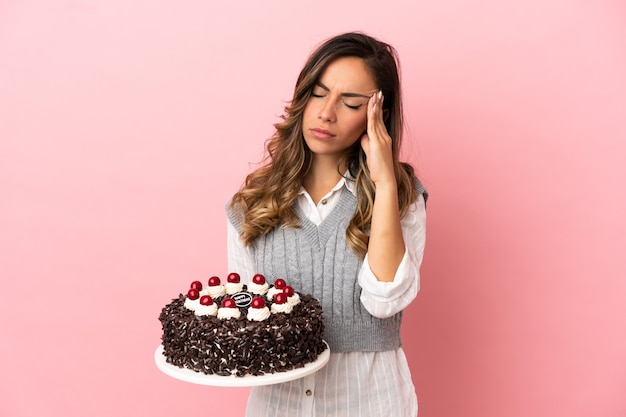 Young woman holding birthday cake over isolated pink background with headache