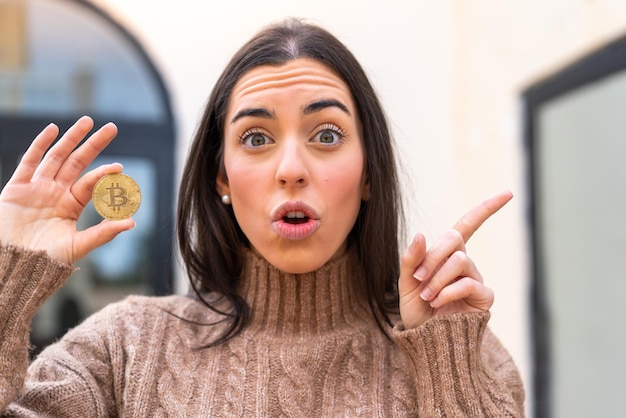 Young woman holding a Bitcoin at outdoors surprised and pointing side