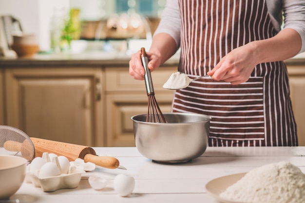 Giovane donna che tiene la ciotola con pasta e frusta, primo piano. una donna con un grembiule a righe sta cucinando in cucina