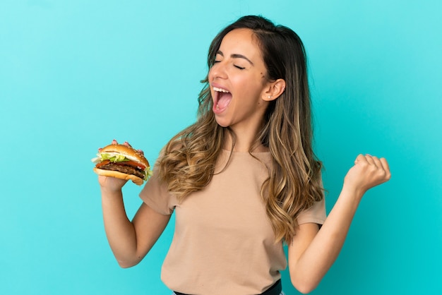 Young woman holding a burger over isolated background celebrating a victory