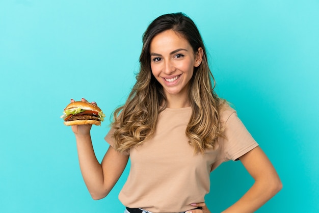 Молодая женщина, держащая гамбургер на изолированном фоне, позирует с руками на бедрах и улыбается