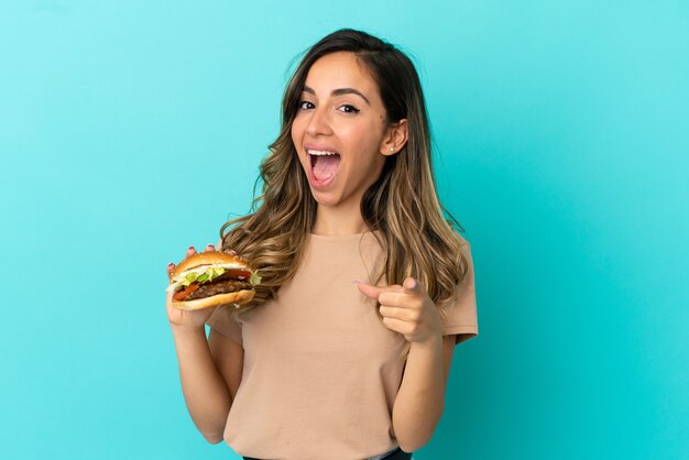 Young woman holding a burger over isolated background surprised and pointing front