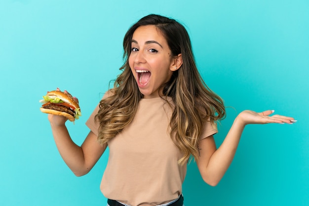 Young woman holding a burger over isolated background with shocked facial expression