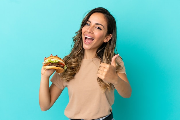 Foto giovane donna che tiene un hamburger su sfondo isolato con il pollice in alto perché è successo qualcosa di buono