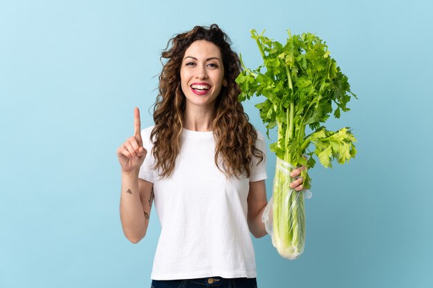 Young woman holding a celery isolated on blue background pointing up a great idea