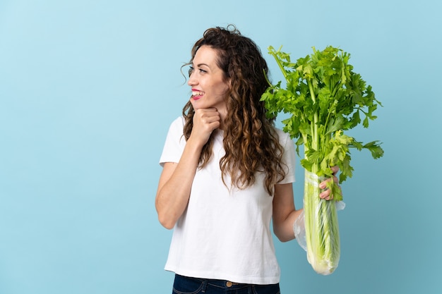 Young woman holding a celery isolated on blue wall thinking an idea and looking side