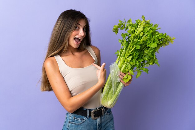 Young woman holding a celery isolated on purple wall with surprise facial expression