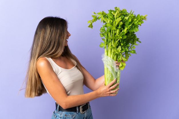 Young woman holding a celery isolated on purple with happy expression