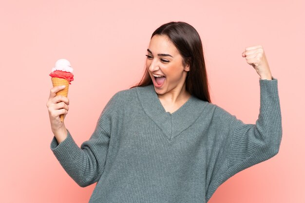 勝利を祝うピンクの背景に分離されたコルネットアイスクリームを保持している若い女性