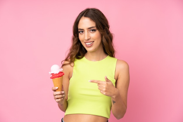 Young woman holding a cornet ice cream over isolated on a pink wall and pointing it