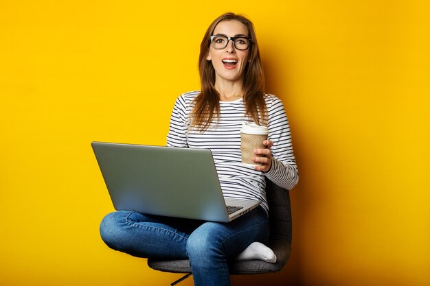 Young woman holding a cup of coffee, sitting on a chair, working on a laptop on an isolated yellow.