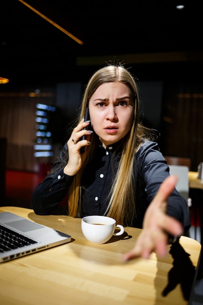 一杯のコーヒーを保持し、電話で話しているラップトップコンピューターを使用して若い女性。在宅勤務の実業家。在宅勤務