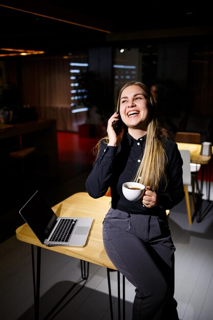 一杯のコーヒーを保持し、電話で話しているラップトップコンピューターを使用して若い女性。自宅で働く実業家。在宅勤務