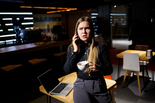 커피 한 잔을 들고 전화 통화를 하는 랩톱 컴퓨터를 사용하는 젊은 여성. 집에서 일하는 사업가. 재택근무