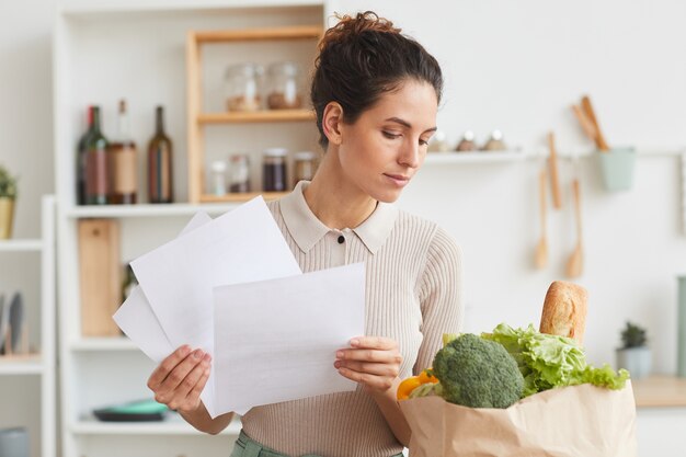 Молодая женщина держит документы и проверяет продукты в бумажном пакете, стоя на кухне