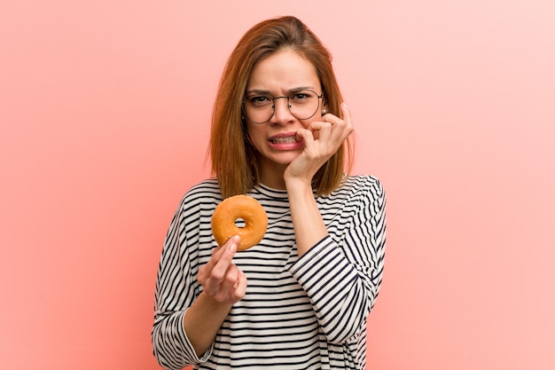 Foto giovane donna con in mano una ciambella che morde le unghie, nervosa e molto ansiosa.