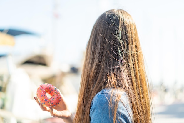 Young woman holding a donut at outdoors in back position