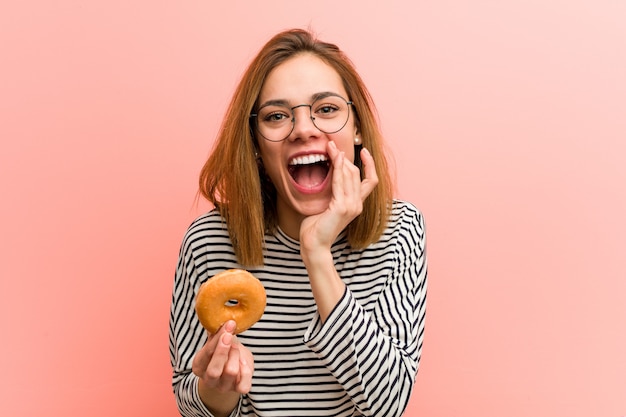 Young woman holding a donut shouting excited to front.