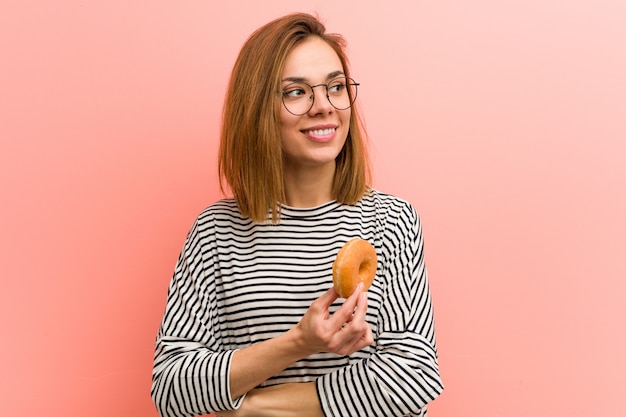 Young woman holding a donut smiling confident with crossed arms.