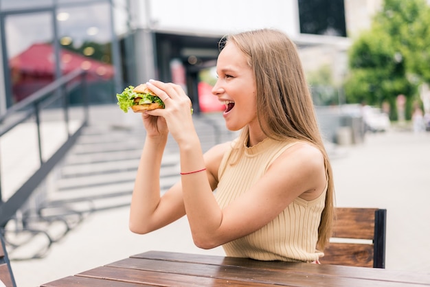 カフェで外でおいしい焼きハンバーガーを食べて保持し、食べる若い女性