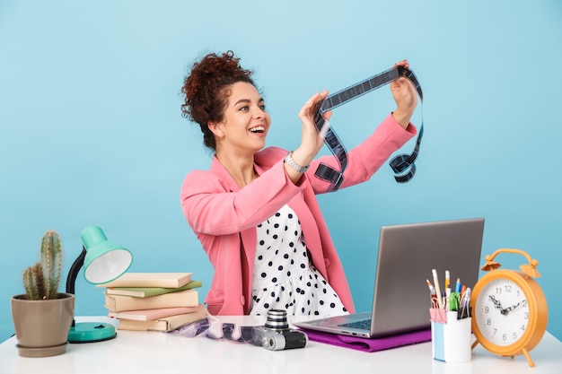 Photo young woman holding footage and smiling while working at desk isolated over blue wall