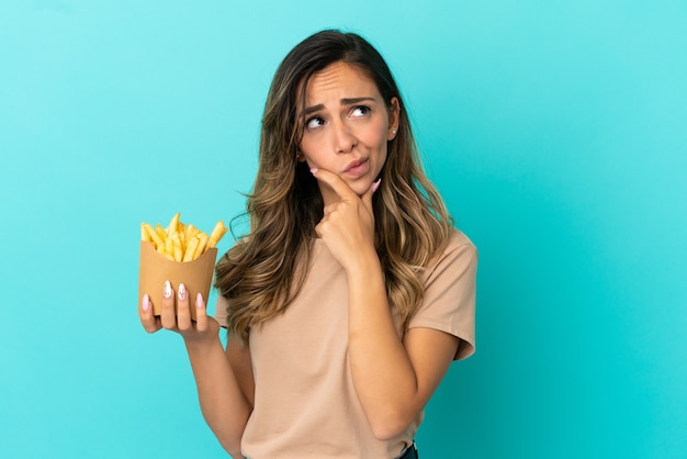 Giovane donna che tiene patatine fritte su sfondo isolato avendo dubbi e pensando