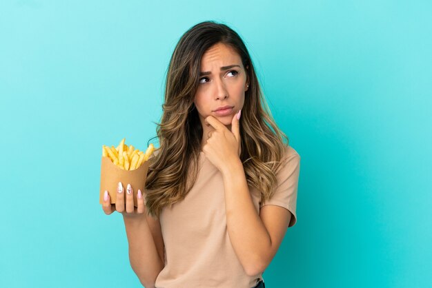 Young woman holding fried chips over isolated background having doubts