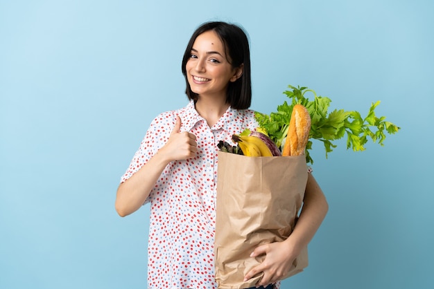 親指を立てるジェスチャーを与える食料品の買い物袋を保持している若い女性