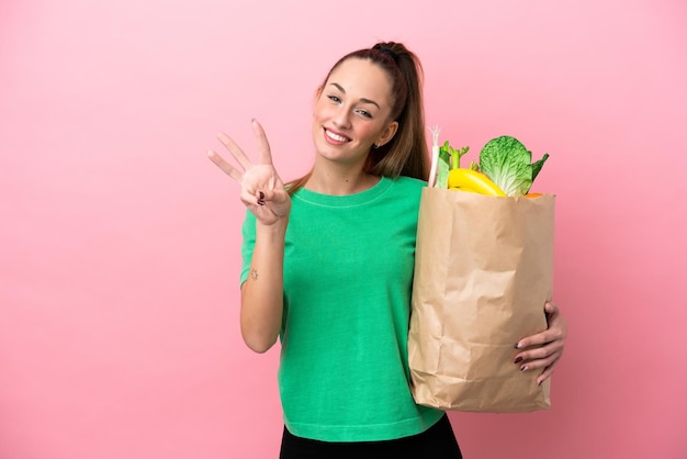 Молодая женщина, держащая продуктовую сумку, счастлива и считает три пальцами