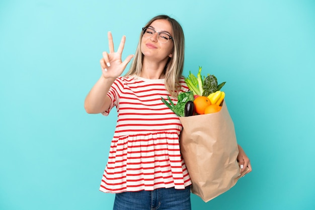 幸せな青い背景で隔離の食料品の買い物袋を保持し、指で3を数える若い女性