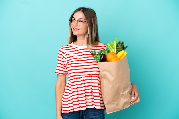 Young woman holding a grocery shopping bag isolated on blue background looking to the side and smiling