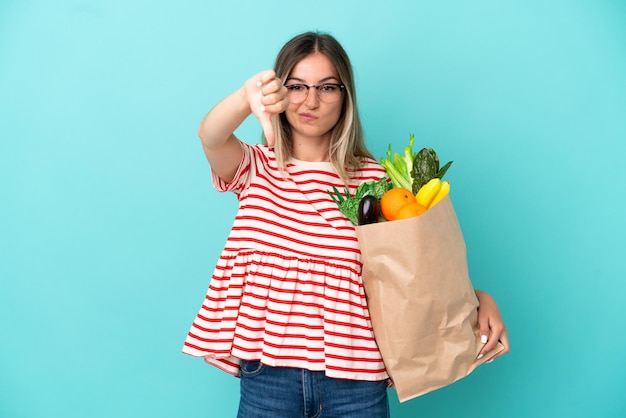 Молодая женщина, держащая продуктовую сумку на синем фоне, показывает большой палец вниз с негативным выражением лица