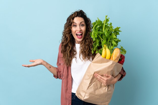 ショックを受けた表情で青い壁に分離された食料品の買い物袋を保持している若い女性