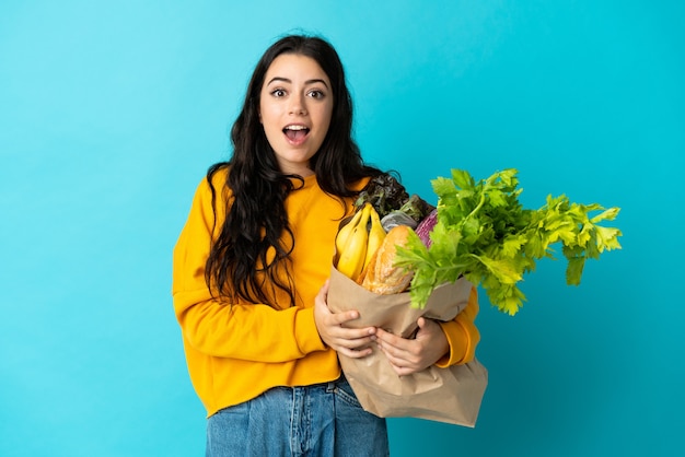 Young woman holding a grocery shopping bag isolated on blue with surprise facial expression