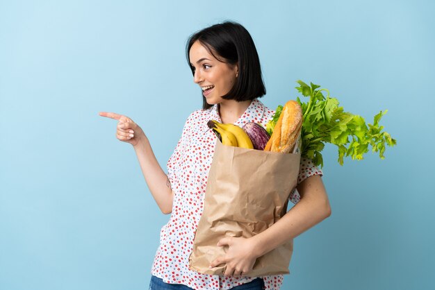 Young woman holding a grocery shopping bag pointing finger to the side and presenting a product