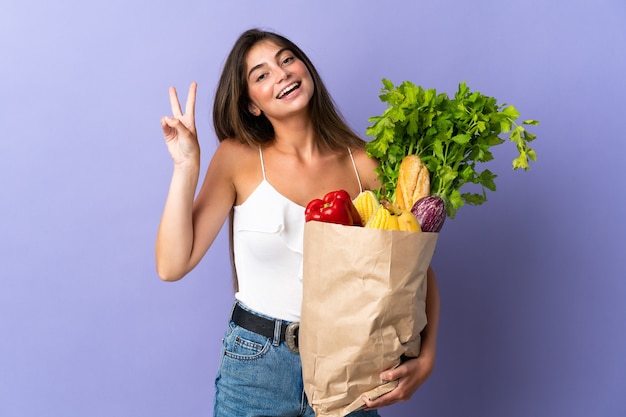 笑顔で勝利のサインを示す食料品の買い物袋を持っている若い女性