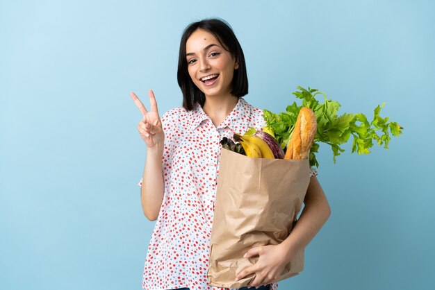 笑顔で勝利のサインを示す食料品の買い物袋を保持している若い女性