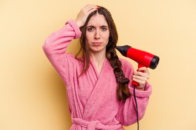 ショックを受けた黄色の背景に分離されたヘアドライヤーを持っている若い女性、彼女は重要な会議を思い出しました。